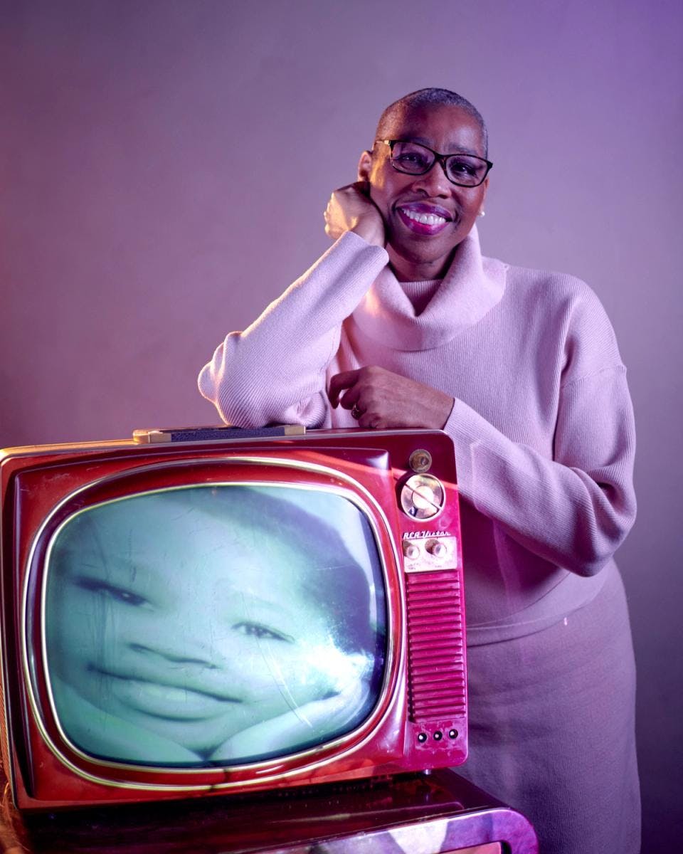A woman smiles and leans against an old TV that has the face of a young girl on it