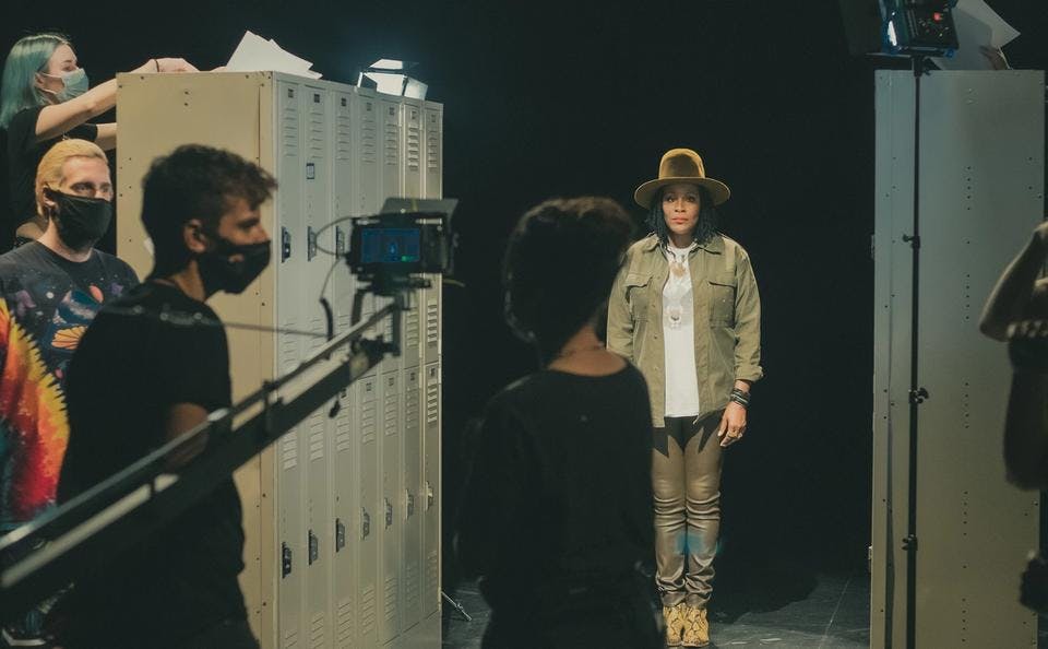 A woman stands on a music video set with lockers, with cameras visible