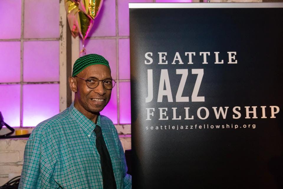 A man smiles standing next to a poster that says "Seattle Jazz Fellowship"